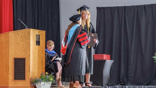 Hannah Brett receives award from Jennifer McConville on stage in caps and gowns on stage receiving diplomas, with Tiffany Heng-Moss member nearby.