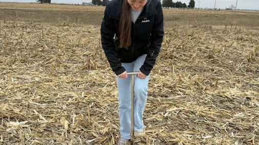 Person in a field using a soil sampling probe under cloudy skies.