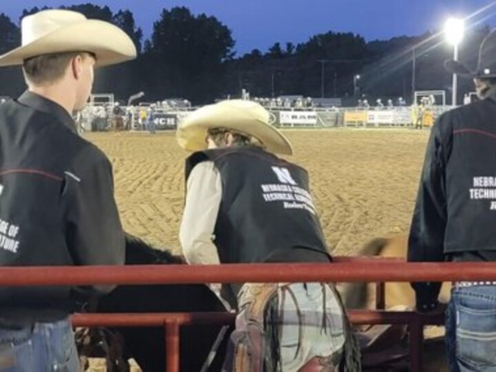 Rodeo team members in cowboy hats and vests at an evening rodeo arena with sandy ground.