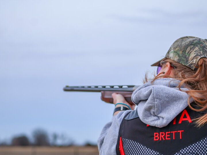 Person in camouflage cap aiming a shotgun outdoors under a cloudy sky.