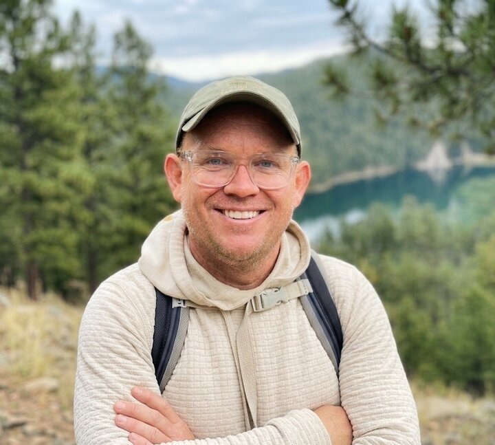 Kurt Elder, NCTA Alumni President, standing and smiling with his arms crossed with a mountain side background